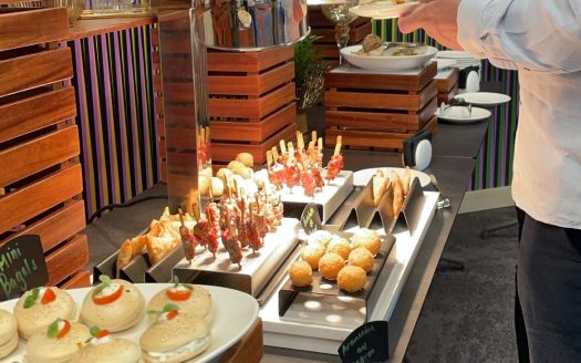 Une personne se servant à un buffet avec une variété de plats, notamment des mini-burgers, des brochettes et d'autres entrées, présentés avec élégance dans un cadre moderne à l'Expo de Dubaï 2020.