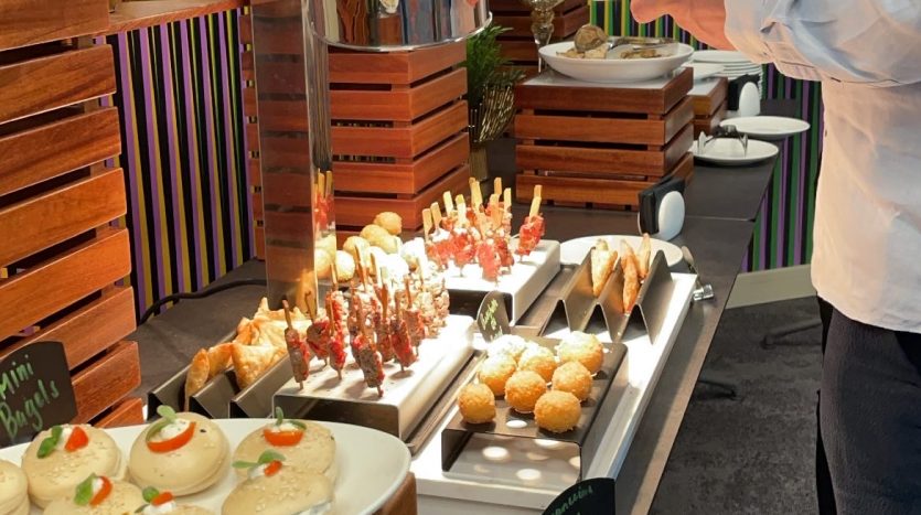 Une personne se servant à un buffet avec une variété de plats, notamment des mini-burgers, des brochettes et d&#039;autres entrées, présentés avec élégance dans un cadre moderne à l&#039;Expo de Dubaï 2020.