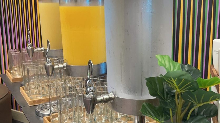 Deux distributeurs de boissons, l&#039;un avec du jus d&#039;orange et l&#039;autre avec de l&#039;eau, sont installés sur une table avec des verres à côté d&#039;eux, sur un fond de mur à rayures colorées décoré de plantes en pot au Dubaï