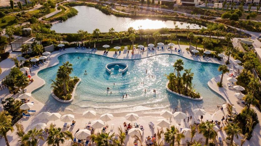 Vue aérienne d&#039;une grande piscine de style complexe à Damas Hills 3, entourée de palmiers et de chaises longues blanches, avec des gens nageant et se relaxant au bord de l&#039;eau, reflétant le coucher du soleil.