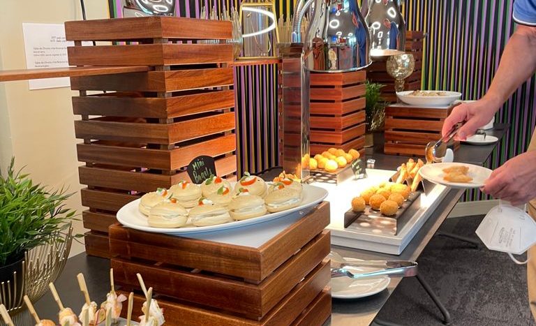 Un homme sert de la nourriture lors d&#039;un buffet coloré dans une luxueuse villa de Dubaï, avec une gamme d&#039;entrées et de desserts présentés sur des plats de service modernes, sur un fond à rayures verticales multicolores.