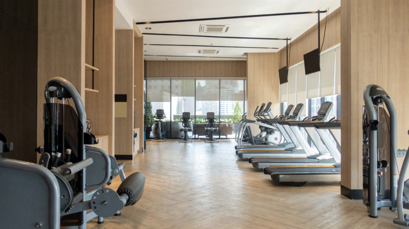 Intérieur d&#039;une salle de sport moderne avec des équipements d&#039;exercice comprenant des tapis roulants et des appareils de musculation, du parquet, de grandes fenêtres et une vue sur la ville de Binghatti Dubaï.