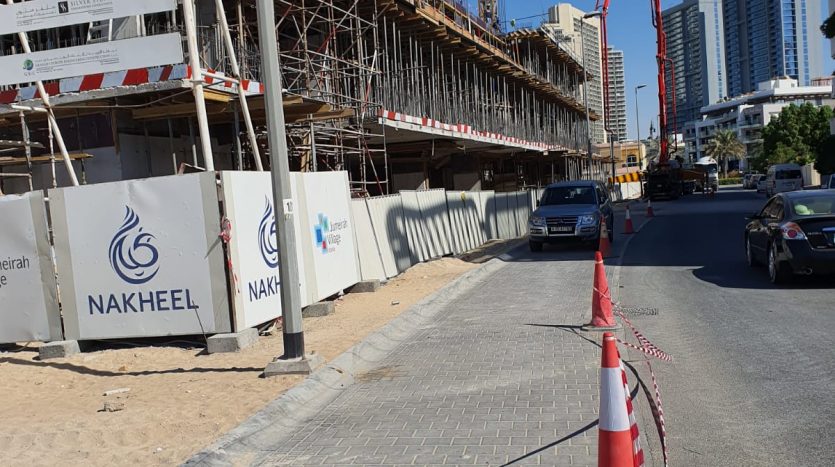 Un chantier de construction dans une rue urbaine de Binghatti Dubaï avec un échafaudage, une grue et une pompe à ciment, marqués par des cônes de sécurité, sous un ciel clair.