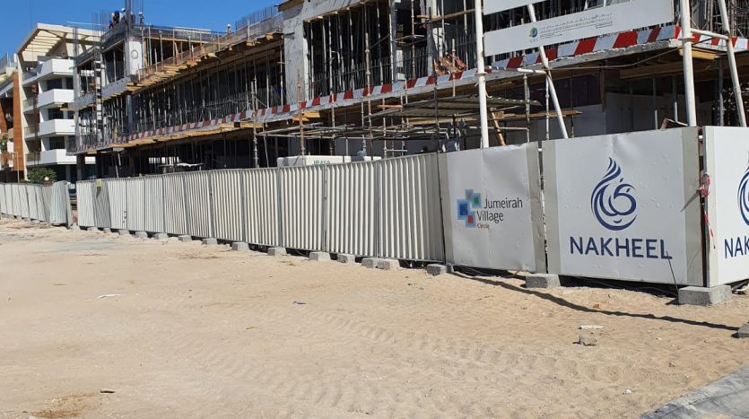 Un chantier de construction avec un grand bâtiment en cours de développement par Binghatti Dubaï, entouré d&#039;une clôture métallique et comportant diverses signalisations d&#039;entreprise. Un ciel bleu clair et un premier plan sablonneux sont visibles.