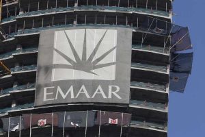 Une grande banderole avec le logo et le nom « Emaar Dubai » est accrochée sur le côté d'un bâtiment partiellement construit avec des sols exposés et des drapeaux flottants.