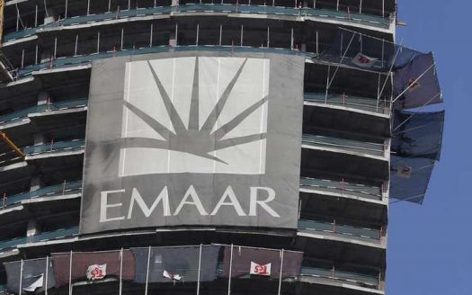 Une grande banderole avec le logo et le nom « Emaar Dubai » est accrochée sur le côté d'un bâtiment partiellement construit avec des sols exposés et des drapeaux flottants.