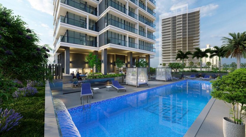 Un immeuble résidentiel moderne de grande hauteur avec une grande piscine extérieure entourée d'un aménagement paysager luxuriant et de chaises longues sous un ciel dégagé de Dubaï.