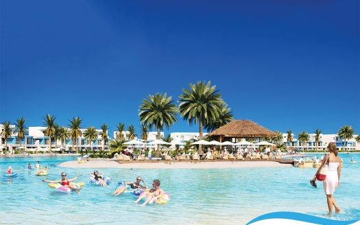 Scène de villégiature tropicale à Portofino, Damac Hills avec une piscine bleu clair, des touristes sur des flotteurs, des palmiers et un ciel lumineux. Des bâtiments blancs bordent l’arrière-plan. Une femme