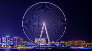 Une grande roue illuminée domine l'horizon nocturne, entourée de bâtiments illuminés à Dubaï, reflétant des couleurs vibrantes sur l'eau devant elle.