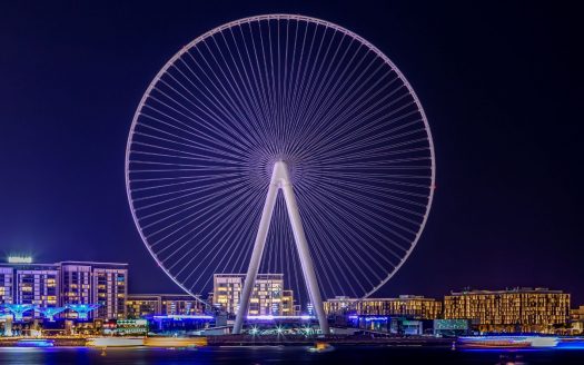 Une grande roue illuminée domine l'horizon nocturne, entourée de bâtiments illuminés à Dubaï, reflétant des couleurs vibrantes sur l'eau devant elle.