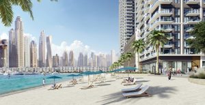Un lungomare soleggiato con sedie a sdraio sotto ombrelloni blu, accanto a un moderno edificio Emaar, con lo skyline della città sullo sfondo e persone che passeggiano tranquillamente sulla spiaggia.