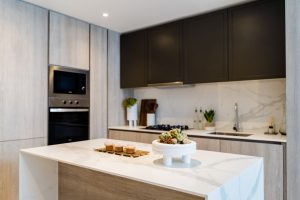 Cuisine moderne avec armoires supérieures sombres et armoires inférieures en bois clair, îlot de comptoir en marbre blanc, appareils électroménagers intégrés en acier inoxydable et plantes décoratives Oxford 212.