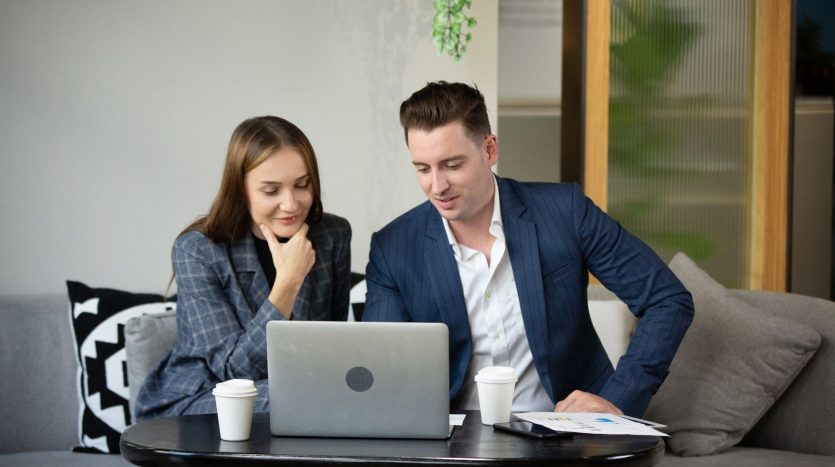 Deux professionnels, une femme et un homme, sont assis à une table et examinent quelque chose sur un ordinateur portable. ils sourient et discutent, avec des tasses à café et des documents sur la table.