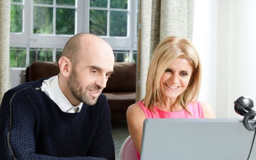 Un homme et une femme souriant et regardant ensemble l’écran d’un ordinateur portable dans une pièce lumineuse dotée de grandes fenêtres. l'homme est chauve et vêtu d'un pull, tandis que la femme a les cheveux blonds et porte un haut rose.