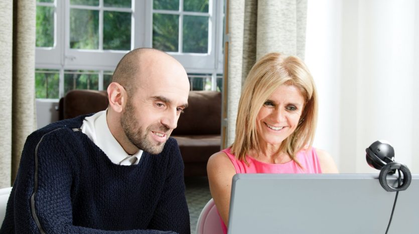 Un homme et une femme souriant et regardant ensemble l’écran d’un ordinateur portable dans une pièce lumineuse dotée de grandes fenêtres. l&#039;homme est chauve et vêtu d&#039;un pull, tandis que la femme a les cheveux blonds et porte un haut rose.