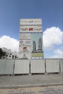 Un grand panneau publicitaire comportant des logos et des textes sur l’investissement à Dubaï se dresse devant un chantier de construction avec un ciel bleu clair au-dessus. La clôture au premier plan entoure le développement en cours.