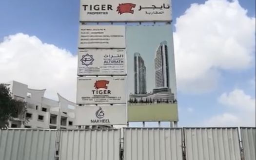 Un grand panneau publicitaire comportant des logos et des textes sur l’investissement à Dubaï se dresse devant un chantier de construction avec un ciel bleu clair au-dessus. La clôture au premier plan entoure le développement en cours.