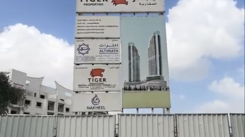 Un grand panneau publicitaire comportant des logos et des textes sur l’investissement à Dubaï se dresse devant un chantier de construction avec un ciel bleu clair au-dessus. La clôture au premier plan entoure le développement en cours.