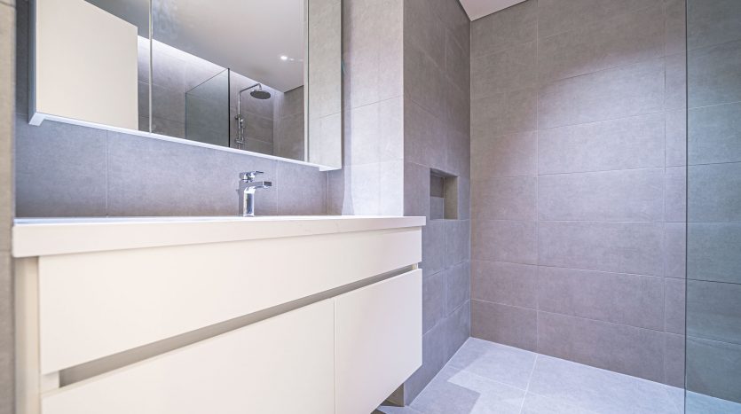 Une salle de bains moderne dans un appartement de Dubaï avec des carreaux gris clair, comprenant une vasque blanche avec un lavabo, un grand miroir et une étagère dissimulée. L&#039;espace est bien éclairé et dispose d&#039;un espace propre