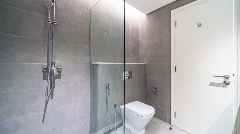 Salle de bains moderne avec cabine de douche en verre, système de douche mural, carrelage gris, toilettes blanches et porte sur le côté droit dans une villa de Dubaï.