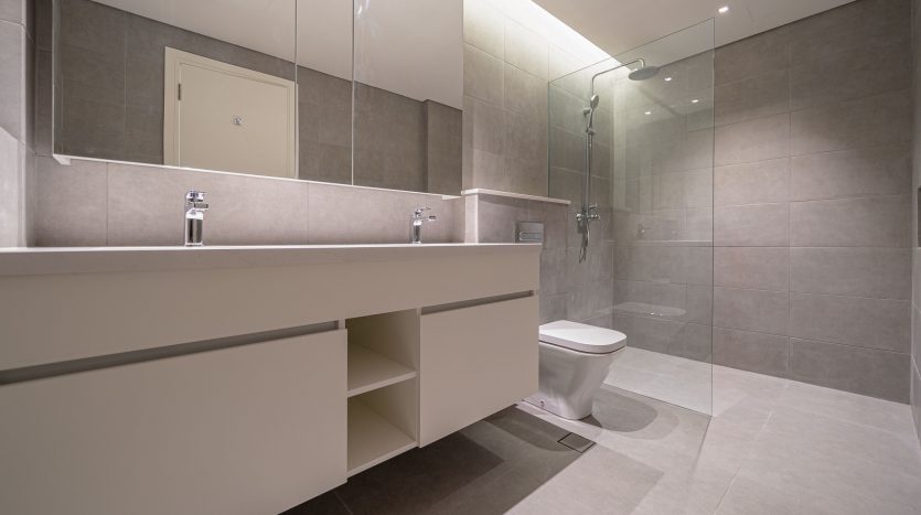 Une salle de bains moderne dans une villa de Dubaï comprenant un grand miroir, une vanité flottante blanche avec des étagères ouvertes, des toilettes murales et une douche vitrée, le tout sur des murs carrelés gris