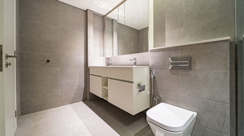Une salle de bains moderne présente des murs et un sol en carrelage gris avec une vanité blanche, un miroir et des toilettes sur la droite. Il y a aussi une porte en bois partiellement visible sur la gauche dans cette villa Dubaï.