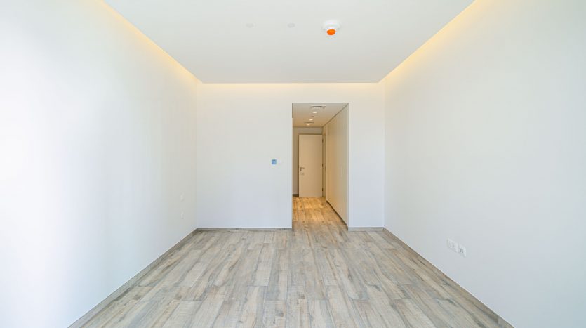 Un couloir vide et étroit dans un appartement de Dubaï avec du parquet en bois clair, des murs blancs et une porte au fond sous un plafonnier lumineux.