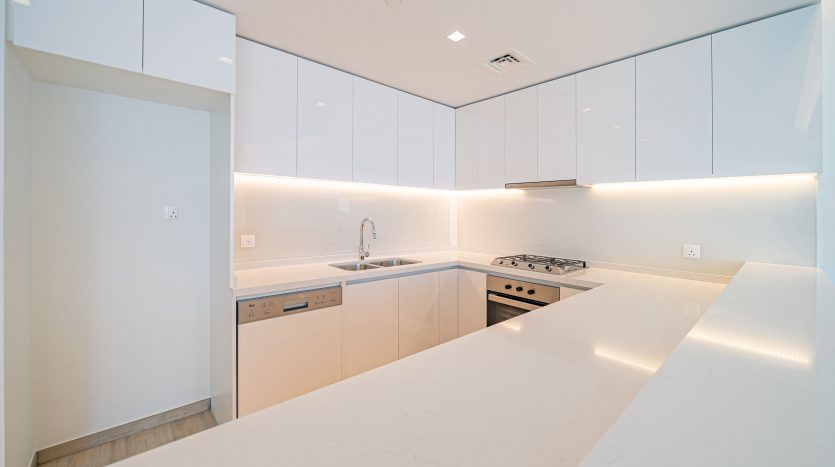 Une cuisine moderne et lumineuse avec des armoires et des comptoirs blancs, un éclairage sous les armoires, une cuisinière intégrée et un design minimaliste pour attirer ceux qui cherchent à investir à Dubaï.