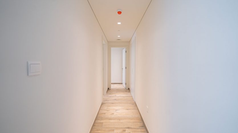 Un couloir long et étroit dans un immeuble moderne aux murs blancs et au parquet, menant à une porte fermée dans un appartement à Dubaï. Un seul détecteur de fumée rouge est monté au plafond.