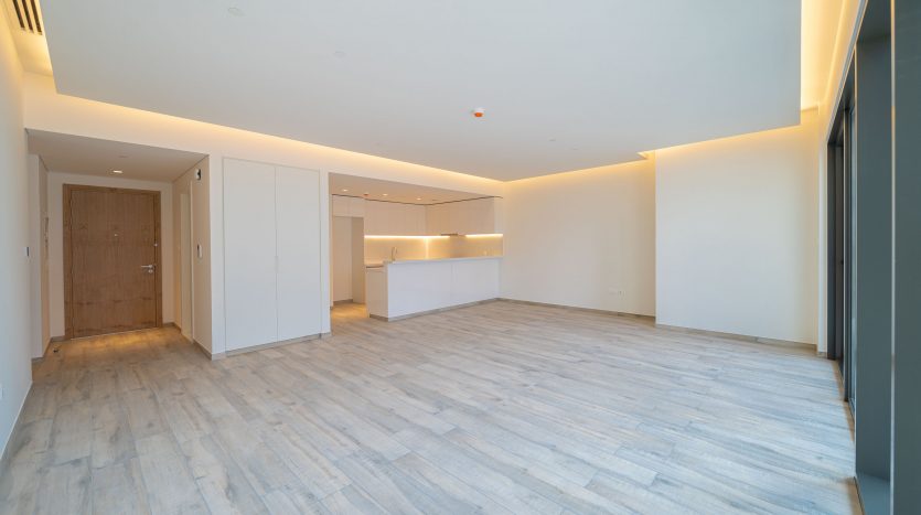 Une pièce vide spacieuse et moderne avec du parquet en bois clair, des murs blancs et un éclairage indirect au plafond, comprenant un coin cuisine ouvert avec un îlot blanc dans un appartement recherché de Dubaï.