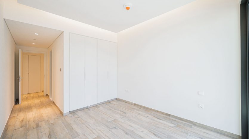 Un couloir spacieux et vide dans un appartement moderne à Dubaï, avec du parquet en bois clair, des murs blancs et des armoires blanches intégrées. Une porte fermée se trouve au fond.