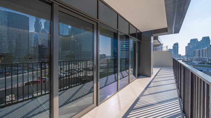 Un balcon moderne dans une villa de Dubaï doté de balustrades en verre transparent offre une vue sur un paysage urbain animé et des routes très fréquentées, sous un ciel dégagé. Le balcon est ombragé par le bâtiment, créant un