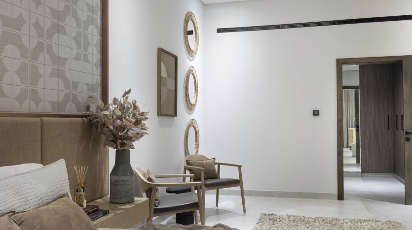 Intérieur de chambre moderne dans une villa de Dubaï comprenant un lit soigné avec une literie taupe, une chaise en bois avec coussin, une petite table d&#039;appoint en bois, des miroirs décoratifs au mur et une plante en pot luxuriante