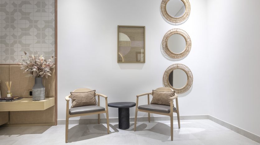 Une salle d&#039;attente moderne dans un appartement de Dubaï avec deux fauteuils en bois avec des coussins beiges, une petite table ronde noire, des miroirs décoratifs au mur et un vase avec des fleurs sur une armoire en bois