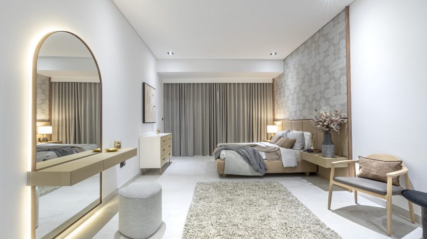 Une chambre moderne et spacieuse dans un appartement de Dubaï comprenant un grand lit avec une literie grise, des meubles en bois, un tapis moelleux, un éclairage tamisé et une décoration minimaliste aux tons neutres.