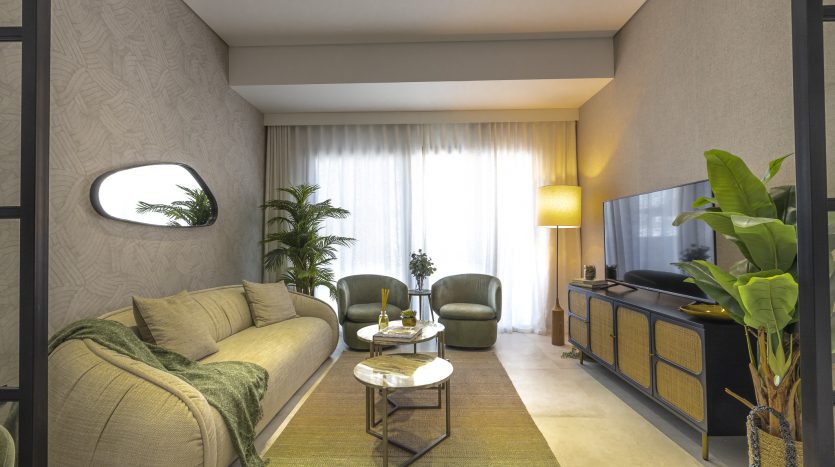 Un salon moderne et confortable dans un appartement de Dubaï avec un canapé beige, deux fauteuils verts, une table basse ronde, une grande télévision, des plantes et un éclairage tamisé provenant de plafonniers cachés et d&#039;un grand