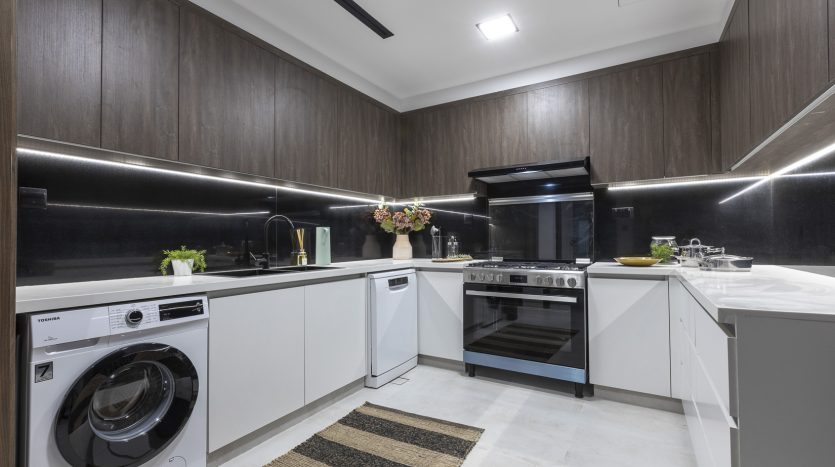Intérieur de cuisine moderne avec armoires en bois sombre, comptoirs blancs et appareils électroménagers intégrés, dont un lave-linge et une cuisinière en acier inoxydable, dans une luxueuse villa à Dubaï. Un bouquet de fleurs ajoute une touche de