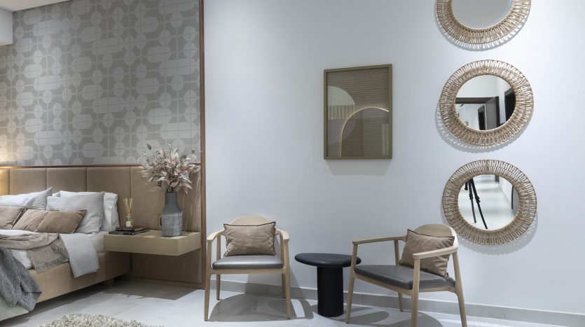 Une chambre moderne dans un appartement de Dubaï avec une palette de couleurs neutres, comprenant un lit double, une tête de lit en bois, deux chaises, une petite table et des miroirs circulaires décoratifs au mur.