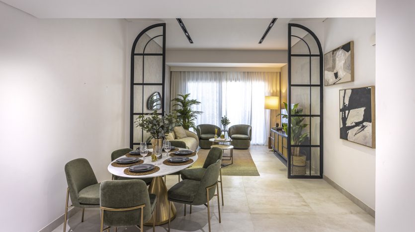 Salle à manger élégante comprenant une table ronde pour six personnes, des chaises en velours vert et des plantes décoratives dans une villa de Dubaï, avec un coin salon confortable visible à travers les arcades.
