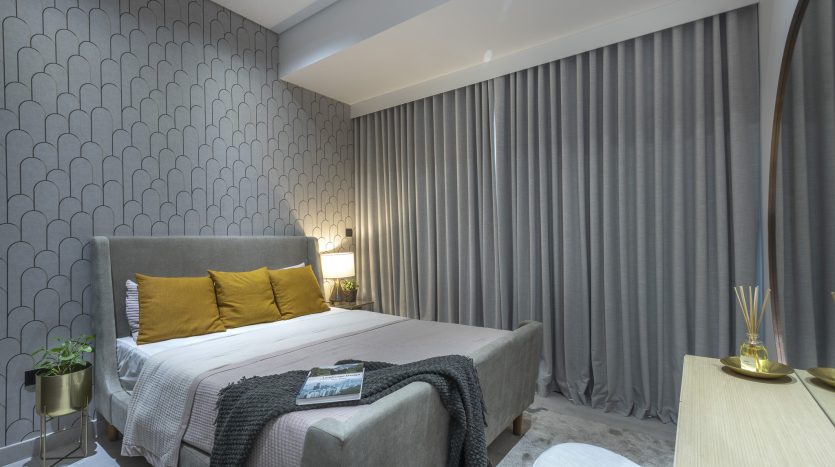 Une chambre moderne dans un appartement de Dubaï avec un grand lit recouvert d&#039;oreillers gris et jaunes, du papier peint texturé, de lourds rideaux gris et un éclairage tamisé. Les objets de décoration comprennent des livres et une petite lampe.