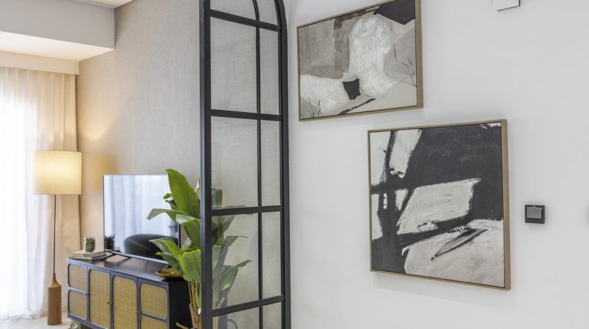 Un salon moderne dans un appartement de Dubaï avec une télévision à écran plat sur un meuble en bois, des œuvres d&#039;art abstraites en noir et blanc sur les murs et une grande plante à côté d&#039;un lampadaire. La pièce est neutre