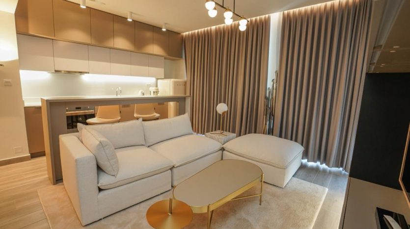 Intérieur de salon moderne avec un canapé blanc et une table basse dorée, des rideaux beiges et une cuisine ouverte avec armoires en arrière-plan d&#039;une villa à Dubaï.