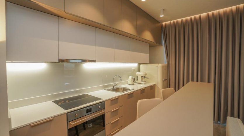 Intérieur de cuisine moderne dans un appartement de Dubaï avec armoires beiges, appareils électroménagers intégrés et éclairage LED sous les armoires suspendues. Un évier et une cuisinière sont visibles avec un rideau drapé à droite