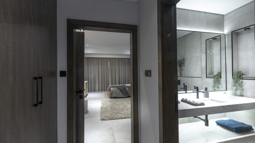 Vue depuis une salle de bains moderne avec deux lavabos et miroirs dans une villa de Dubaï, montrant une porte ouverte menant à une chambre avec un lit et un tapis texturé. Les tons neutres et terreux dominent le décor raffiné