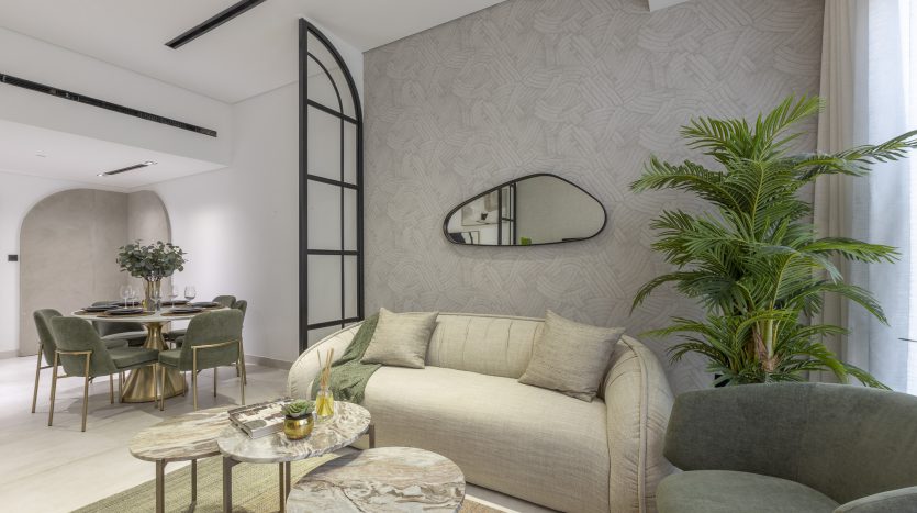 Un salon moderne avec un canapé beige, deux fauteuils verts et une table basse en marbre. Derrière, un espace salle à manger de l&#039;appartement Dubai sous une ouverture cintrée. Grandes plantes et papier peint géométrique