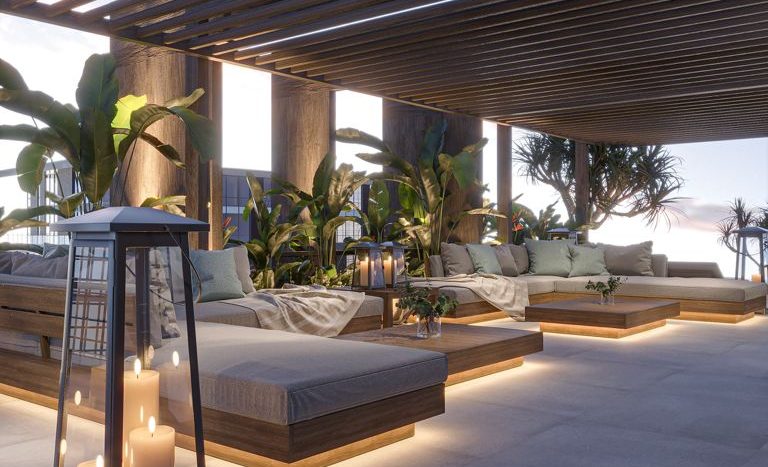 Un coin salon extérieur au crépuscule avec des meubles modernes en bois, des bougies dans des bougeoirs de bon goût et des plantes, sous un élégant auvent en bois dans une villa de Dubaï, surplombant une mer calme.