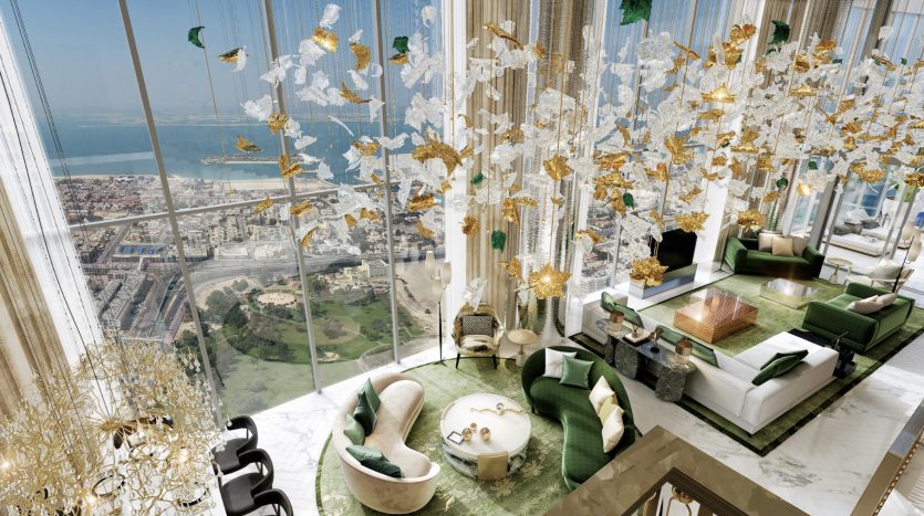 Luxueux salon en hauteur à Dubaï avec des murs en verre offrant une vue panoramique sur la ville et la mer. La pièce présente des décorations opulentes en or et blanc, un mobilier élégant et une palette de couleurs vertes luxuriantes.