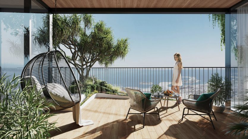 Une femme debout sur un balcon moderne avec parquet, entouré de plantes vertes luxuriantes, surplombant la mer à Dubaï. Le balcon est doté d'un mobilier d'extérieur élégant, notamment d'un fauteuil à œufs suspendu.