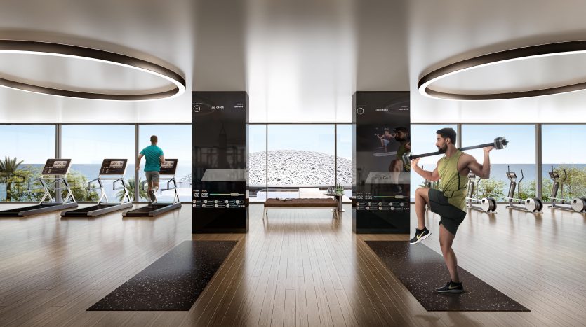 Une salle de sport moderne avec de grandes fenêtres et une vue sur l&#039;océan à Dubaï. Plusieurs appareils d&#039;exercice et deux hommes s&#039;entraînant ; l’un courant sur un tapis roulant et l’autre soulevant des poids.