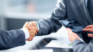 Deux professionnels se serrant la main au-dessus d'un bureau dans une agence immobilière de Dubaï, l'un d'entre eux tenant une tablette numérique et des documents visibles, symbolisant un accord ou un partenariat dans un cadre professionnel.